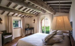 Cozy bedroom featuring rustic wooden beams, a large window with a view, elegant bedding, and a stylish lamp, creating a warm and inviting atmosphere.