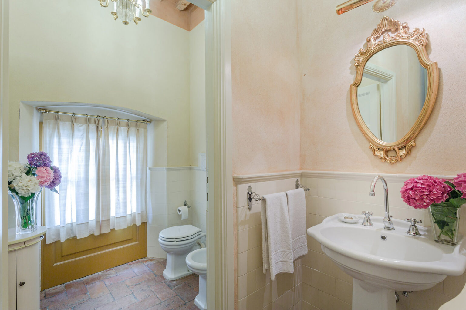 Charming vintage bathroom featuring a white pedestal sink, a decorative mirror, and a cozy window draped with sheer curtains. Fresh flowers add a pop of color, while the soft pastel walls create a warm ambiance. | Only on SopranoVillas