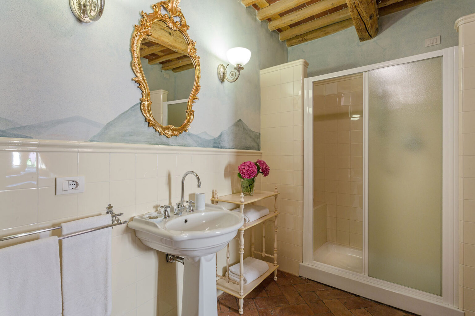 Elegant bathroom featuring a vintage sink, a decorative mirror, and a glass shower. The walls are adorned with soft mountain-themed murals, complemented by a stylish towel rack and fresh flowers, creating a serene atmosphere. | Only on SopranoVillas