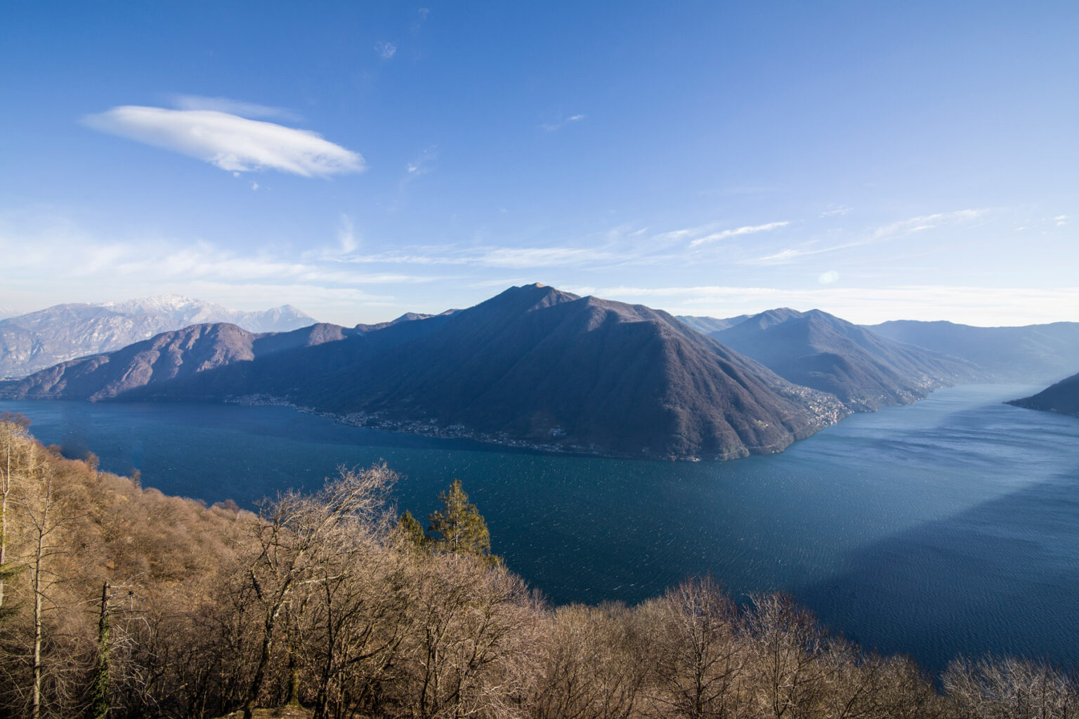 Panoramic view of a serene lake surrounded by mountains under a clear blue sky, showcasing the natural beauty of the landscape. | Only on SopranoVillas