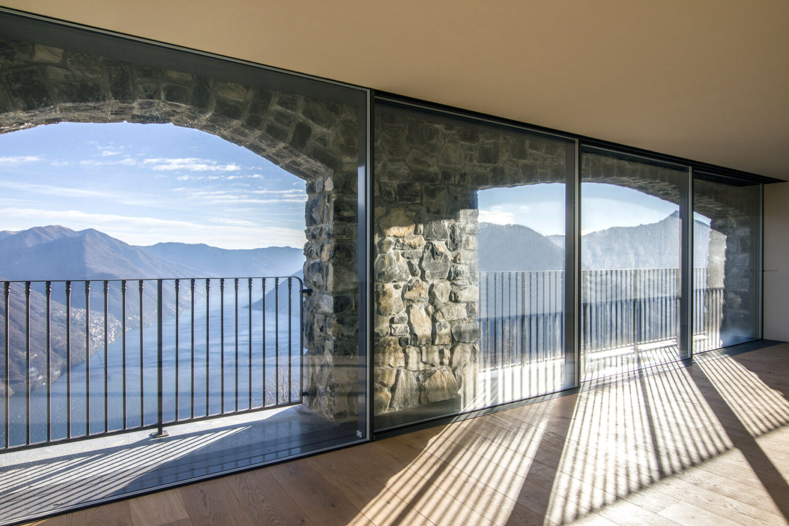 Modern architectural interior featuring large glass windows and stone walls, showcasing a breathtaking view of mountains and a lake, with sunlight casting shadows on the floor. | Only on SopranoVillas
