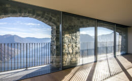Modern architectural interior featuring large glass windows and stone walls, showcasing a breathtaking view of mountains and a lake, with sunlight casting shadows on the floor.