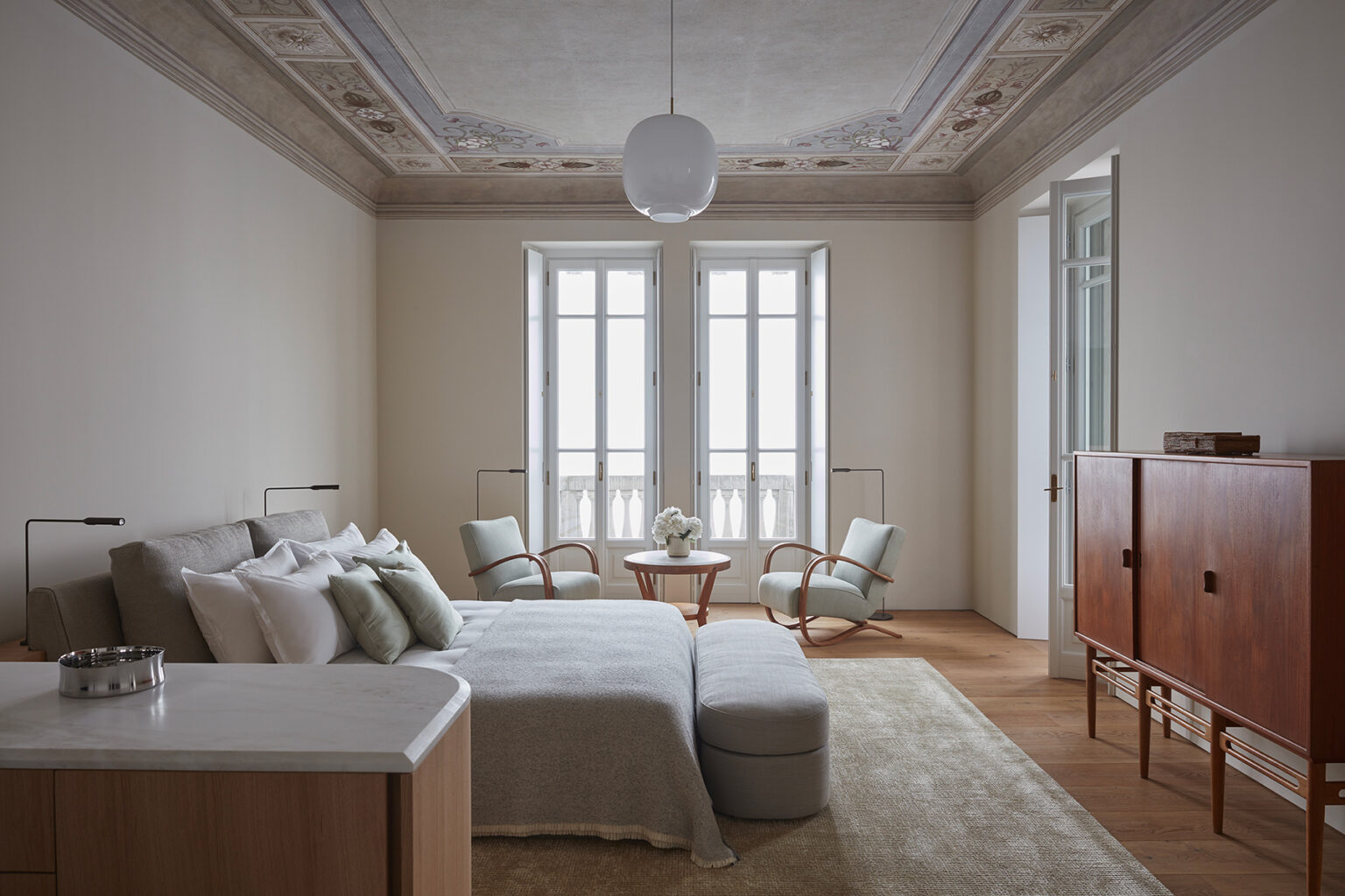 Modern bedroom interior featuring a cozy bed with decorative pillows, two stylish armchairs, a wooden side table, and elegant ceiling details. Natural light floods the space through large windows, enhancing the serene and minimalist design. | Only on SopranoVillas