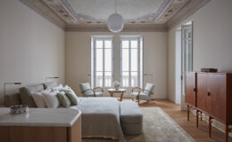 Modern bedroom interior featuring a cozy bed with decorative pillows, two stylish armchairs, a wooden side table, and elegant ceiling details. Natural light floods the space through large windows, enhancing the serene and minimalist design.