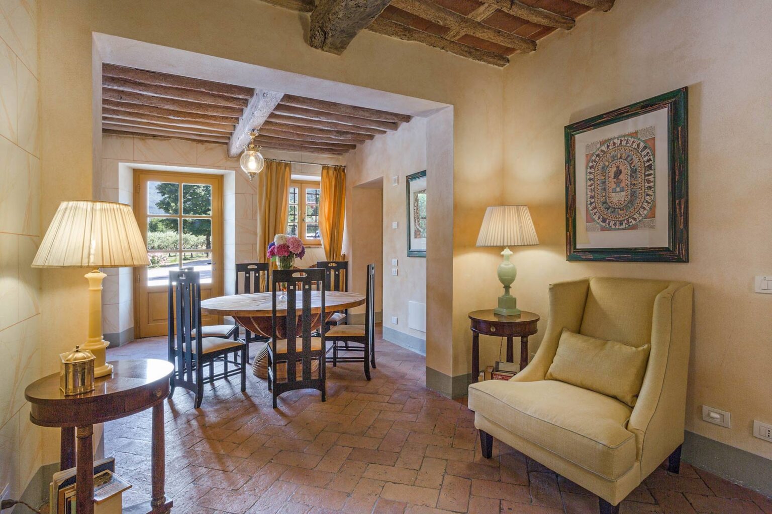 Cozy dining area with a round table and wooden chairs, featuring warm lighting from two lamps, a comfortable yellow armchair, and large windows that invite natural light, set in a rustic interior with exposed wooden beams. | Only on SopranoVillas