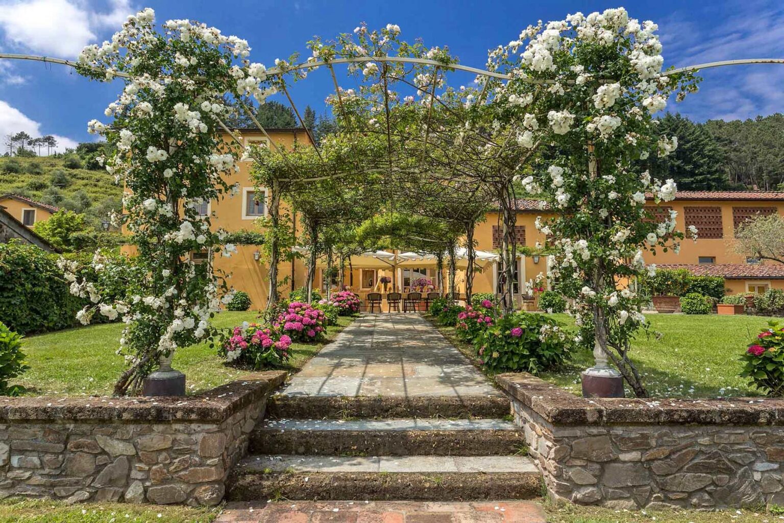 Scenic garden pathway leading to a charming villa, adorned with white roses and colorful flowers, under a clear blue sky. Ideal for outdoor events and relaxation. | Only on SopranoVillas