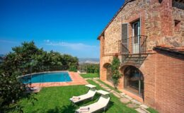 Charming rustic villa with a private pool surrounded by lush greenery and sun loungers, set against a clear blue sky. Ideal for a relaxing getaway in a picturesque landscape.
