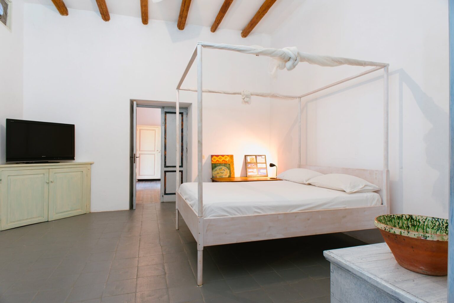 Modern bedroom with a four-poster bed, minimalist decor, and natural light. Features include a flat-screen TV, light-colored furniture, and a potted plant. Ideal for a stylish and comfortable stay. | Only on SopranoVillas
