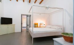 Modern bedroom with a four-poster bed, minimalist decor, and natural light. Features include a flat-screen TV, light-colored furniture, and a potted plant. Ideal for a stylish and comfortable stay.
