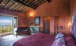 Cozy bedroom with a rustic design, featuring a purple bedspread, wooden beams, and a view of nature through an open door. The room includes a leather sofa and decorative candles, creating a warm and inviting atmosphere.