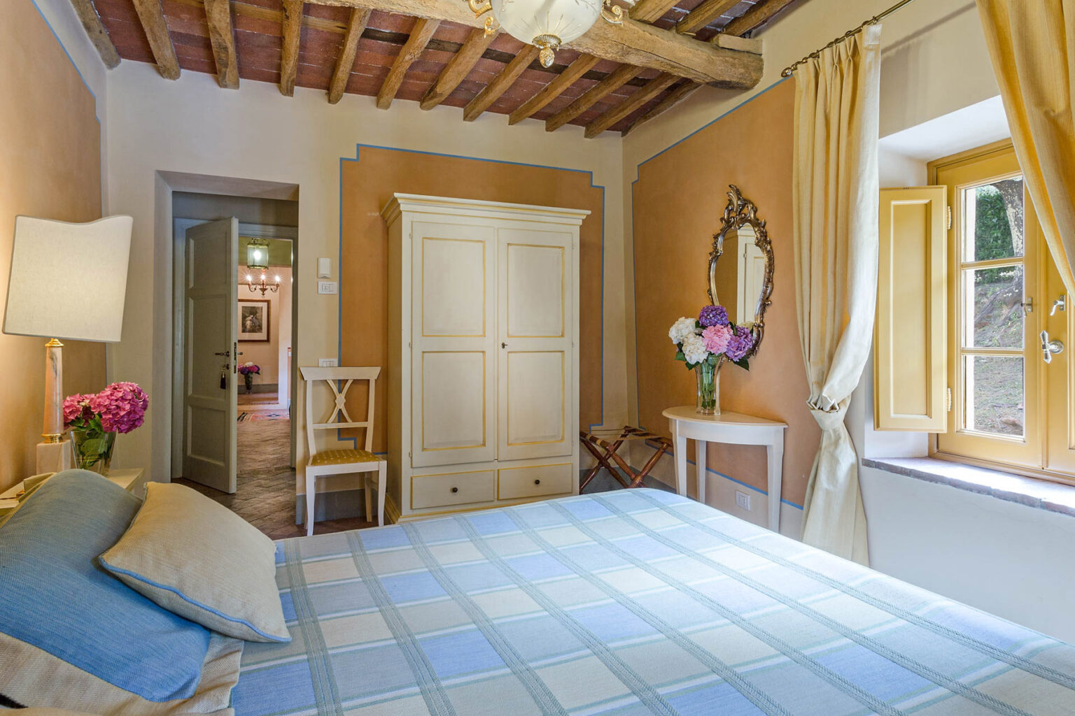 Cozy bedroom featuring a wooden beam ceiling, a comfortable bed with blue and cream bedding, a vintage wardrobe, a small table with a mirror, and a window with yellow curtains letting in natural light. Fresh flowers add a touch of color to the room. | Only on SopranoVillas