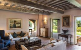 Cozy living room interior featuring rustic wooden beams, comfortable seating, and art on the walls, with large windows offering views of a garden.