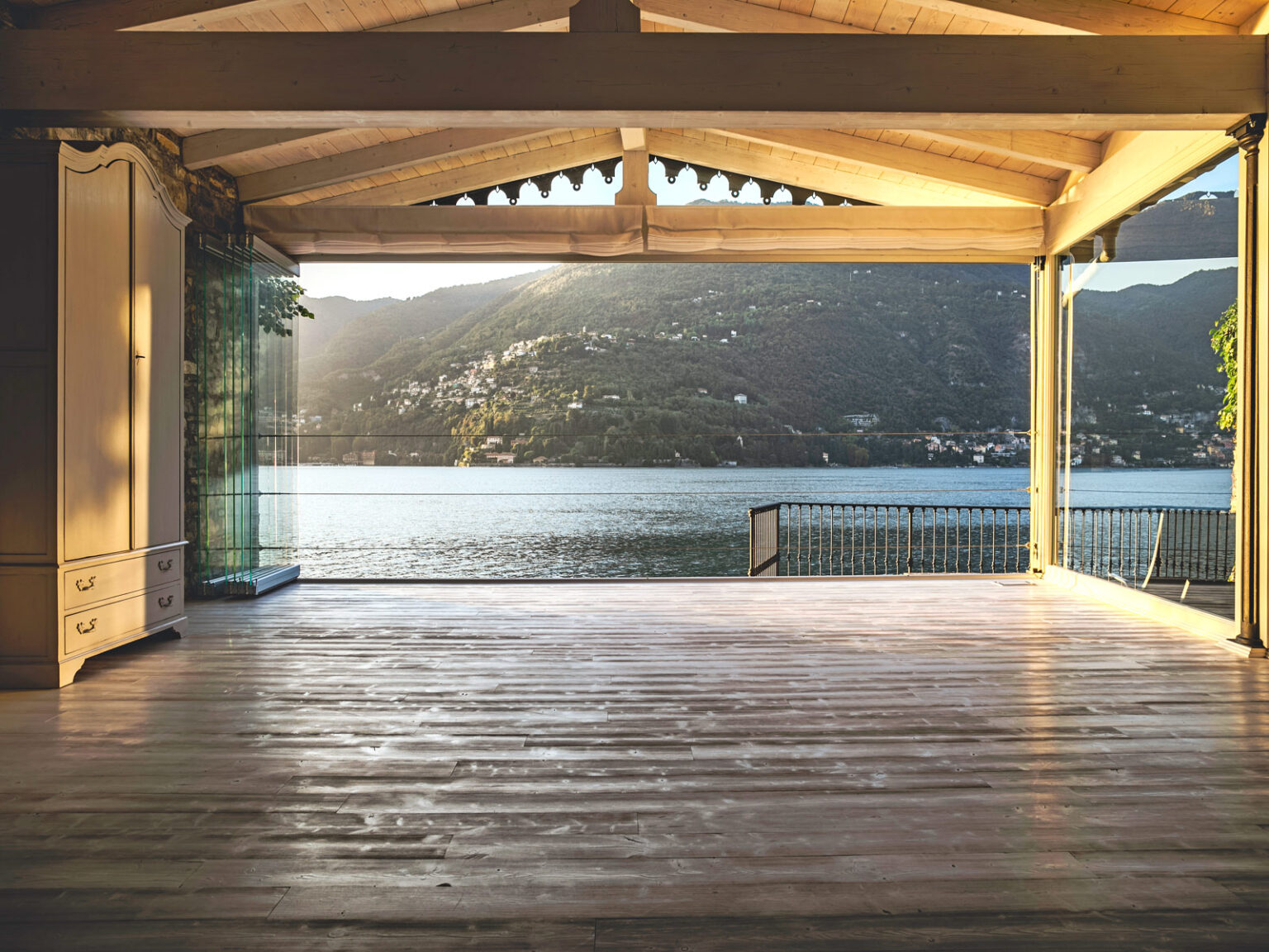 Spacious modern interior with wooden flooring and large glass walls showcasing a stunning view of a lake and mountains during sunset. Ideal for showcasing luxury real estate or vacation homes. | Only on SopranoVillas