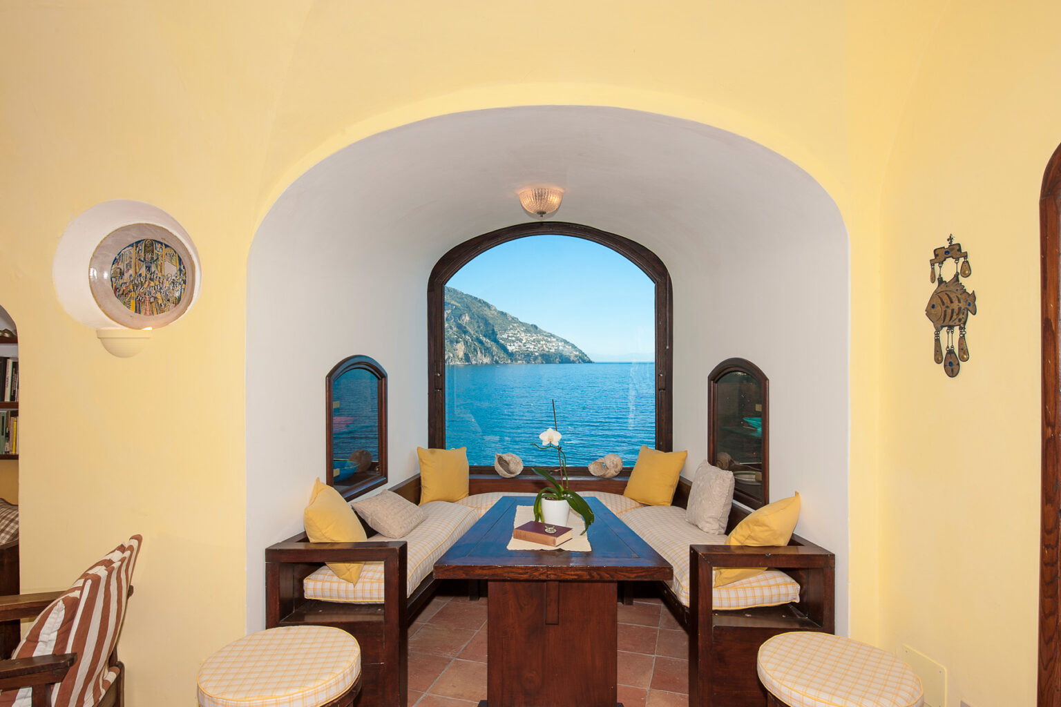 Cozy interior of a seaside living room featuring a large arched window with a scenic view of the ocean and mountains, complemented by comfortable seating and bright yellow walls. | Only on SopranoVillas