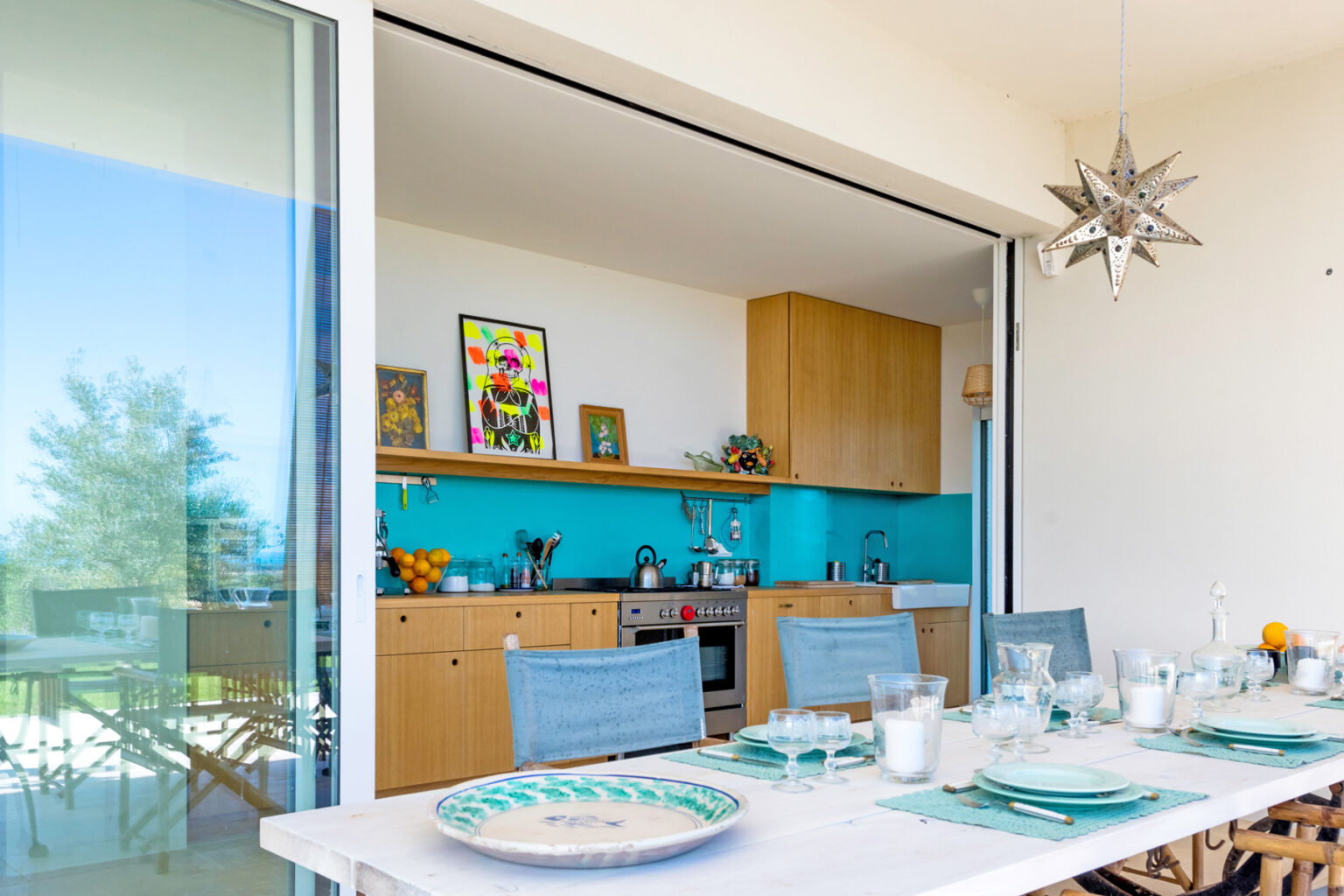Modern dining area with a spacious table set for a meal, featuring a vibrant turquoise kitchen in the background. The design includes wooden cabinetry, decorative artwork, and large sliding glass doors that open to a scenic view. | Only on SopranoVillas