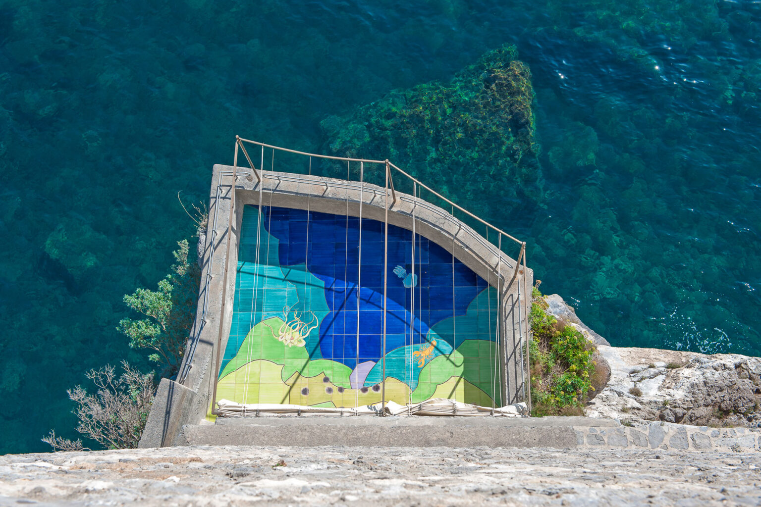 Aerial view of a colorful mural on a concrete platform overlooking clear blue water, featuring vibrant designs and natural elements. | Only on SopranoVillas