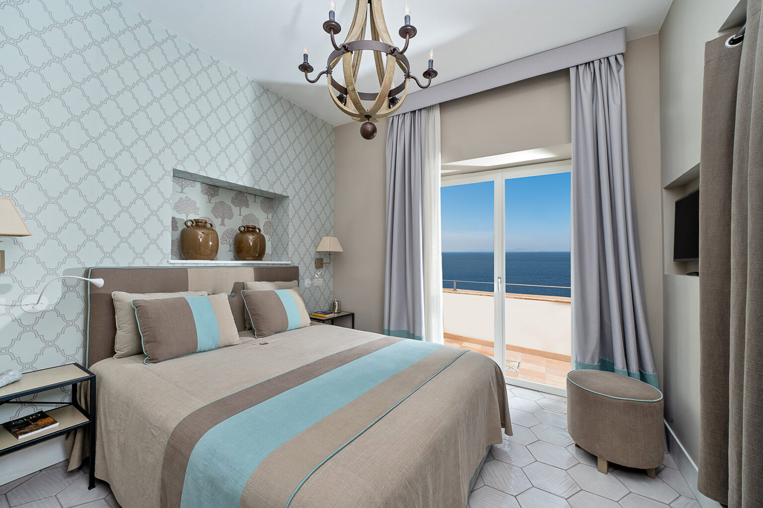 Cozy hotel bedroom with modern decor, featuring a king-sized bed with blue and beige accents, large windows offering a sea view, and elegant lighting fixtures. | Only on SopranoVillas