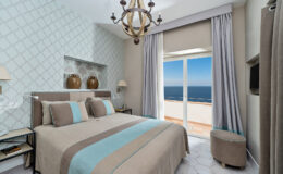 Cozy hotel bedroom with modern decor, featuring a king-sized bed with blue and beige accents, large windows offering a sea view, and elegant lighting fixtures.