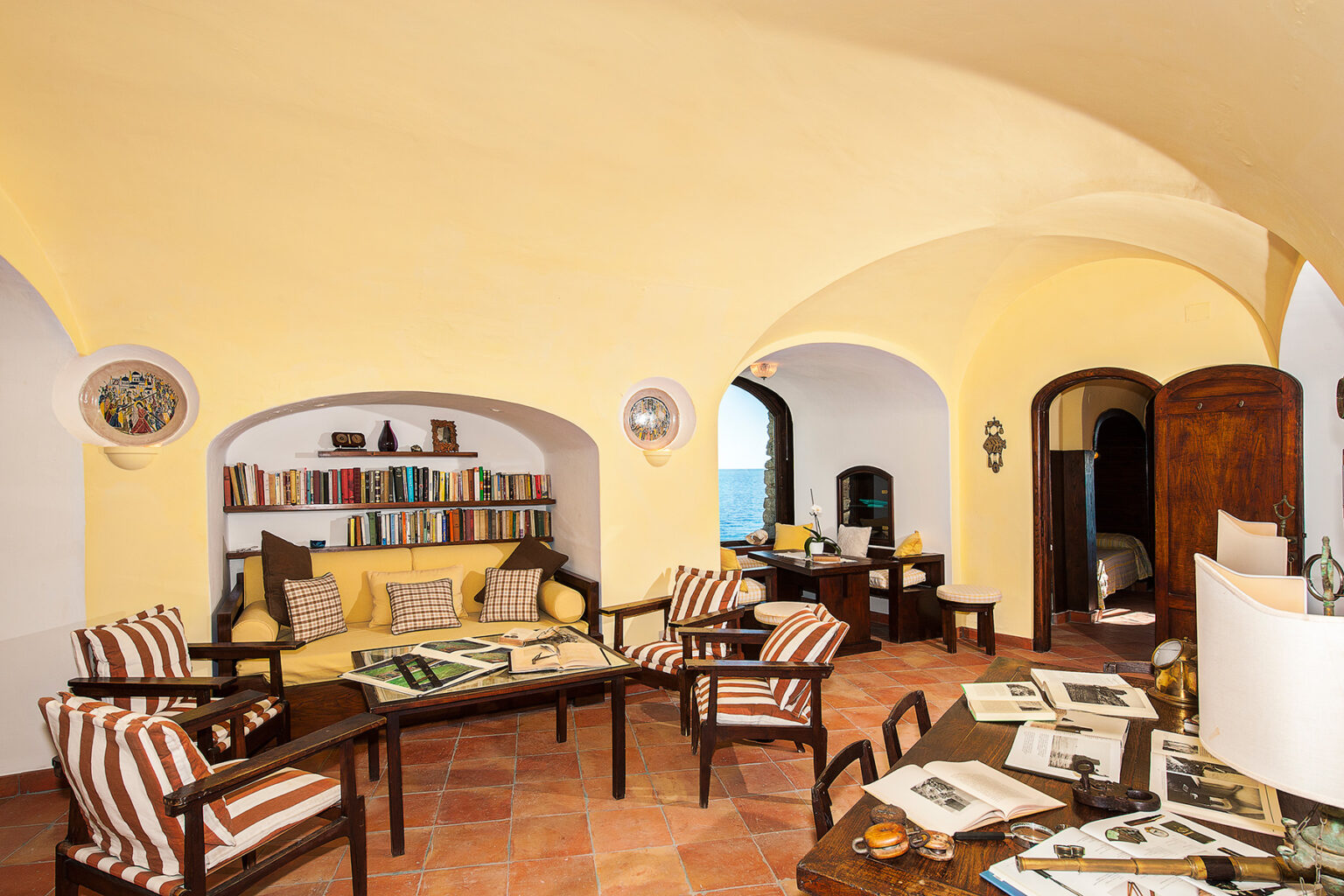 Cozy living room with arched ceilings, yellow walls, and a mix of modern and rustic furniture. Features a comfortable seating area with striped cushions, a wooden table with magazines, and a bookshelf filled with books, creating an inviting atmosphere. | Only on SopranoVillas
