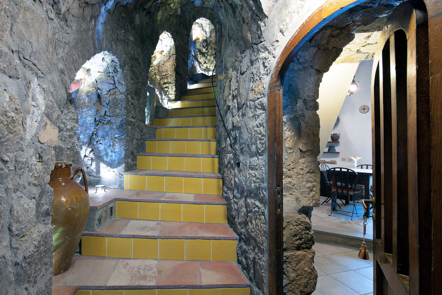 Interior view of a rustic stone staircase leading to a dining area, featuring arched doorways and decorative pottery, illuminated by soft lighting. | Only on SopranoVillas