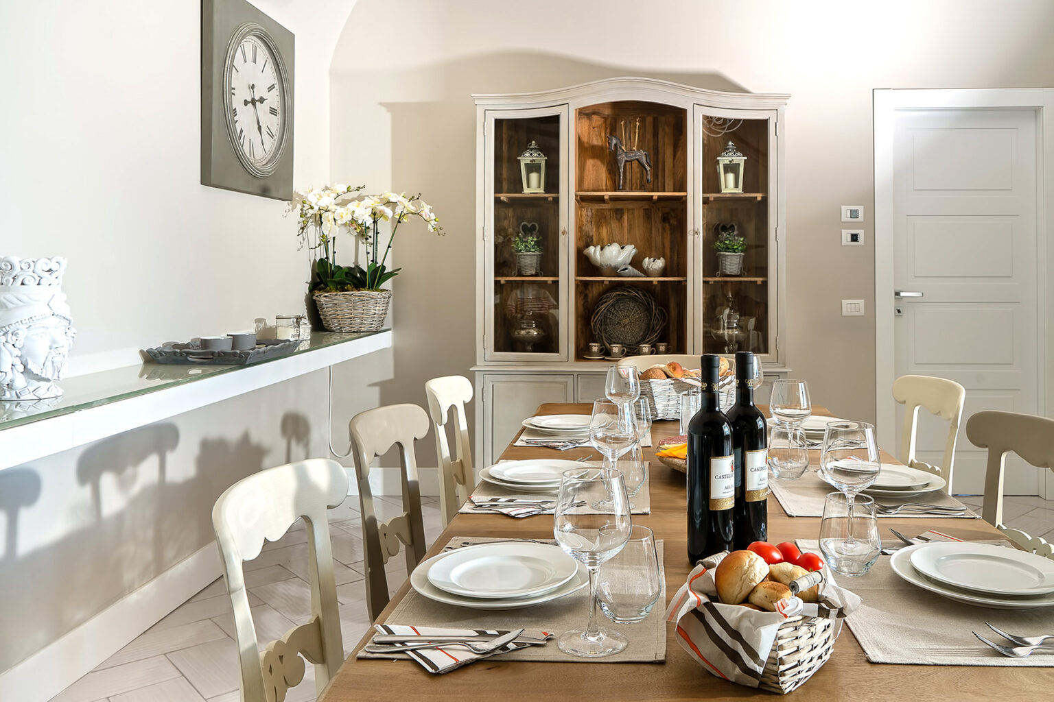 Elegant dining room with a long wooden table set for a meal, featuring wine bottles, glassware, and fresh bread. A light-colored cabinet displays decorative items, while a wall clock and floral arrangements enhance the cozy atmosphere. | Only on SopranoVillas