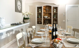 Elegant dining room with a long wooden table set for a meal, featuring wine bottles, glassware, and fresh bread. A light-colored cabinet displays decorative items, while a wall clock and floral arrangements enhance the cozy atmosphere.
