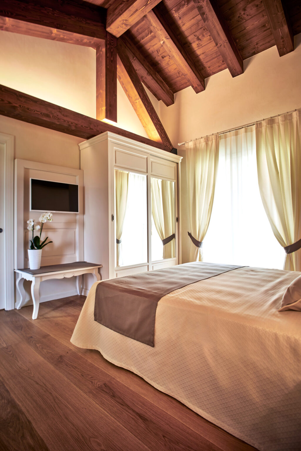 Cozy bedroom interior featuring a neatly made bed with a beige cover, a stylish wardrobe with mirrored doors, a small table, and large windows draped with soft curtains, creating a warm and inviting atmosphere. | Only on SopranoVillas