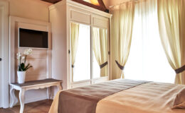 Cozy bedroom interior featuring a neatly made bed with a beige cover, a stylish wardrobe with mirrored doors, a small table, and large windows draped with soft curtains, creating a warm and inviting atmosphere.