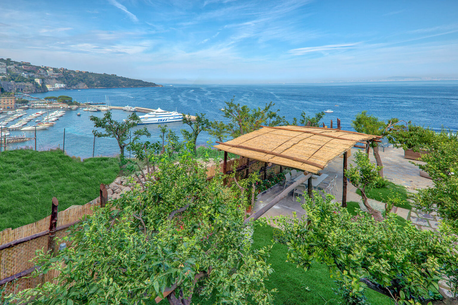 A scenic view of a coastal landscape featuring a wooden pavilion surrounded by lush greenery, overlooking a marina and the ocean under a clear blue sky. | Only on SopranoVillas