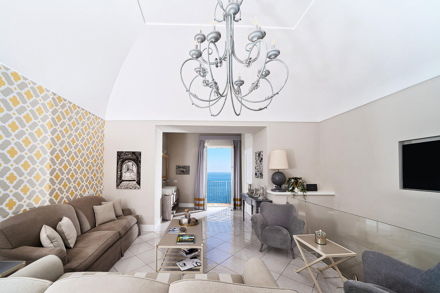 Modern living room with high ceilings, elegant chandelier, and stylish decor. Features a cozy sofa, glass coffee table, and a view of the ocean through large windows. Bright, inviting space with contemporary design elements. | Only on SopranoVillas