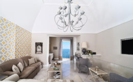 Modern living room with high ceilings, elegant chandelier, and stylish decor. Features a cozy sofa, glass coffee table, and a view of the ocean through large windows. Bright, inviting space with contemporary design elements.
