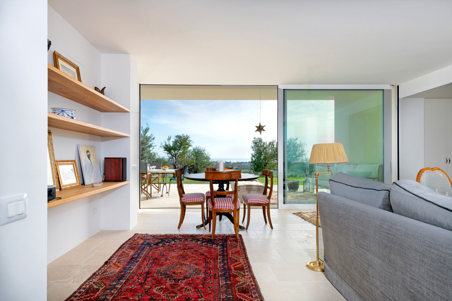 Modern living room interior featuring a cozy seating area with a gray sofa, a traditional rug, and a dining table set for four. Large windows offer a scenic view of greenery, enhancing natural light in the space. Stylish wooden shelves display decorative items and books, contributing to the contemporary aesthetic. | Only on SopranoVillas