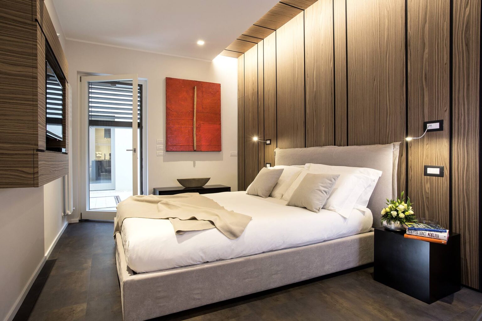 Modern bedroom featuring a minimalist design with a comfortable bed, wooden accent walls, and stylish decor. The room includes bedside tables with lamps and a vibrant piece of abstract art, creating a cozy and inviting atmosphere. | Only on SopranoVillas