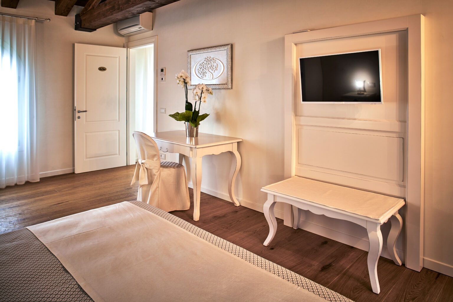 Elegant hotel room featuring a white desk and chair, a flat-screen TV mounted on the wall, and a stylish bench. Natural light streams in through sheer curtains, enhancing the cozy atmosphere. | Only on SopranoVillas