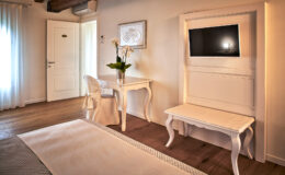 Elegant hotel room featuring a white desk and chair, a flat-screen TV mounted on the wall, and a stylish bench. Natural light streams in through sheer curtains, enhancing the cozy atmosphere.