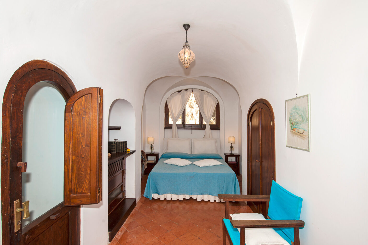 Cozy bedroom featuring a blue bedspread, wooden doors, and charming décor, set in a well-lit, arched room with a vintage touch. | Only on SopranoVillas