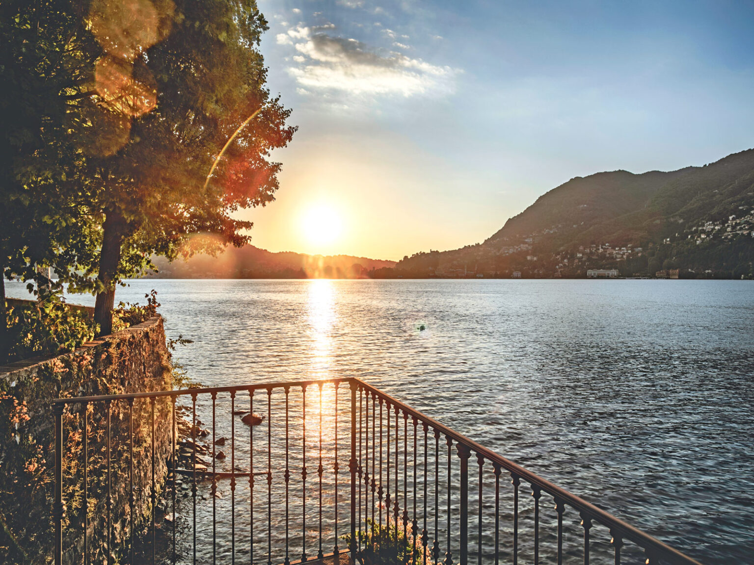 Sunset over a tranquil lake with mountains in the background, featuring a railing and trees along the shore, creating a serene landscape scene. | Only on SopranoVillas