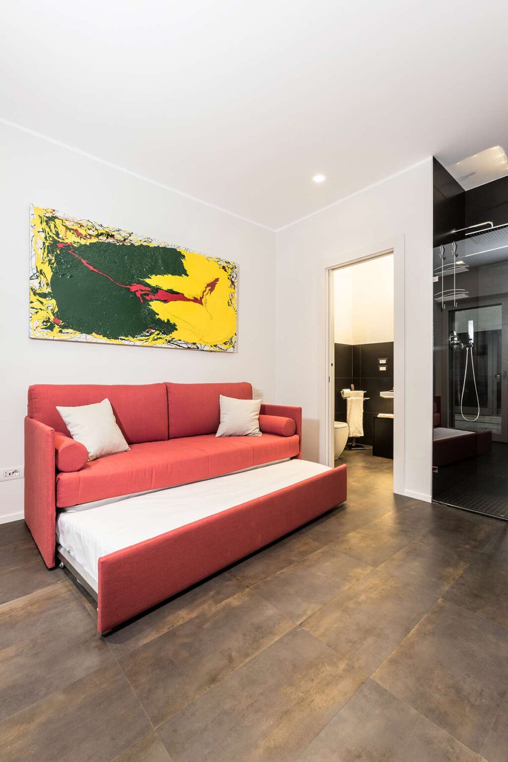 Modern living room featuring a red sofa bed with white cushions, vibrant abstract artwork on the wall, and a doorway leading to a bathroom. The space showcases a contemporary design with a sleek floor and minimalist decor. | Only on SopranoVillas