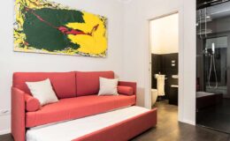 Modern living room featuring a red sofa bed with white cushions, vibrant abstract artwork on the wall, and a doorway leading to a bathroom. The space showcases a contemporary design with a sleek floor and minimalist decor.