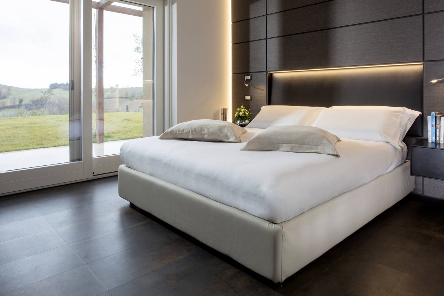 Modern bedroom interior featuring a stylish bed with white linens and decorative pillows, large windows allowing natural light, and a serene outdoor view. Ideal for showcasing contemporary home design and comfort. | Only on SopranoVillas