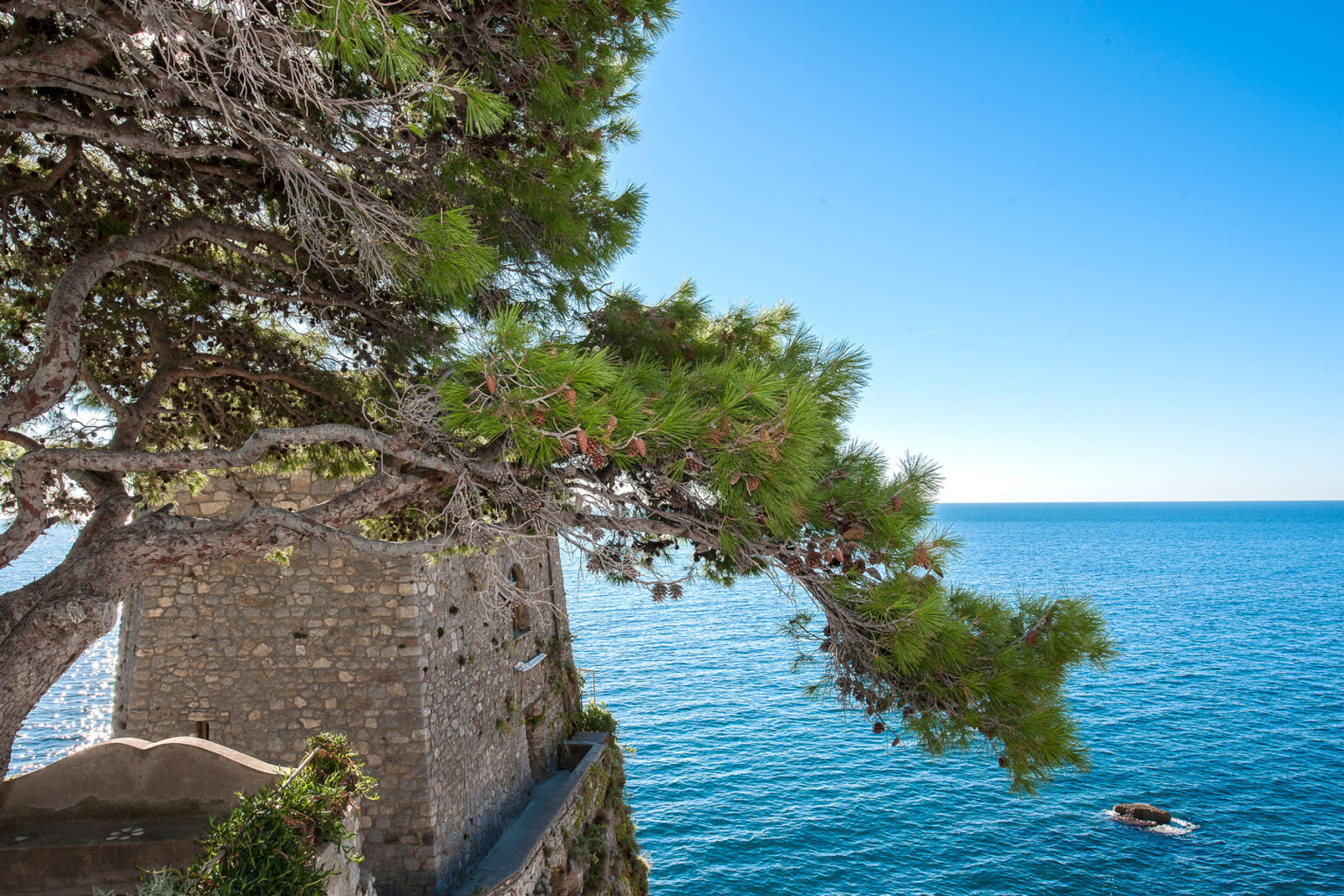 Scenic view of a coastal landscape featuring a stone wall, lush green tree branches, and a tranquil blue sea under a clear sky. | Only on SopranoVillas