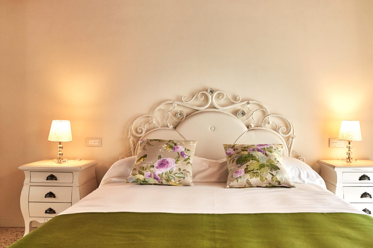 Elegant bedroom with a white ornate headboard, decorative floral pillows, a cozy green throw, and soft bedside lamps creating a warm ambiance. | Only on SopranoVillas