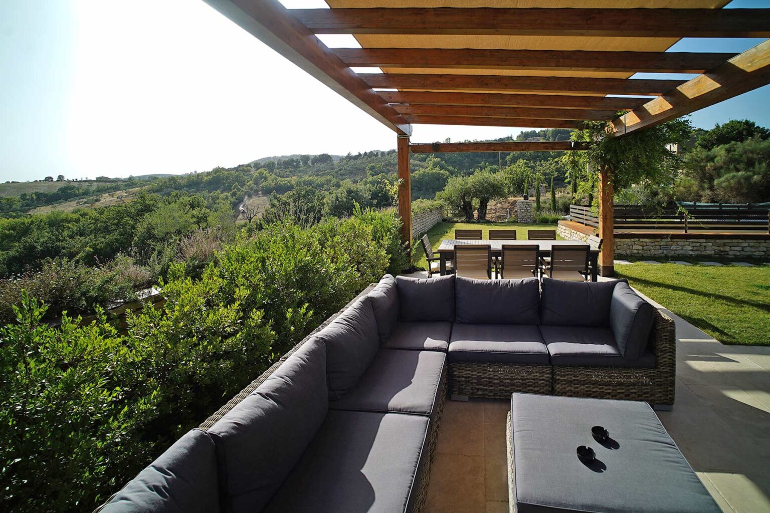 Outdoor seating area with a wooden pergola, featuring a gray sectional sofa and a coffee table, overlooking a lush green landscape and distant hills. Ideal for relaxation and outdoor gatherings. | Only on SopranoVillas