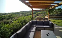 Outdoor seating area with a wooden pergola, featuring a gray sectional sofa and a coffee table, overlooking a lush green landscape and distant hills. Ideal for relaxation and outdoor gatherings.