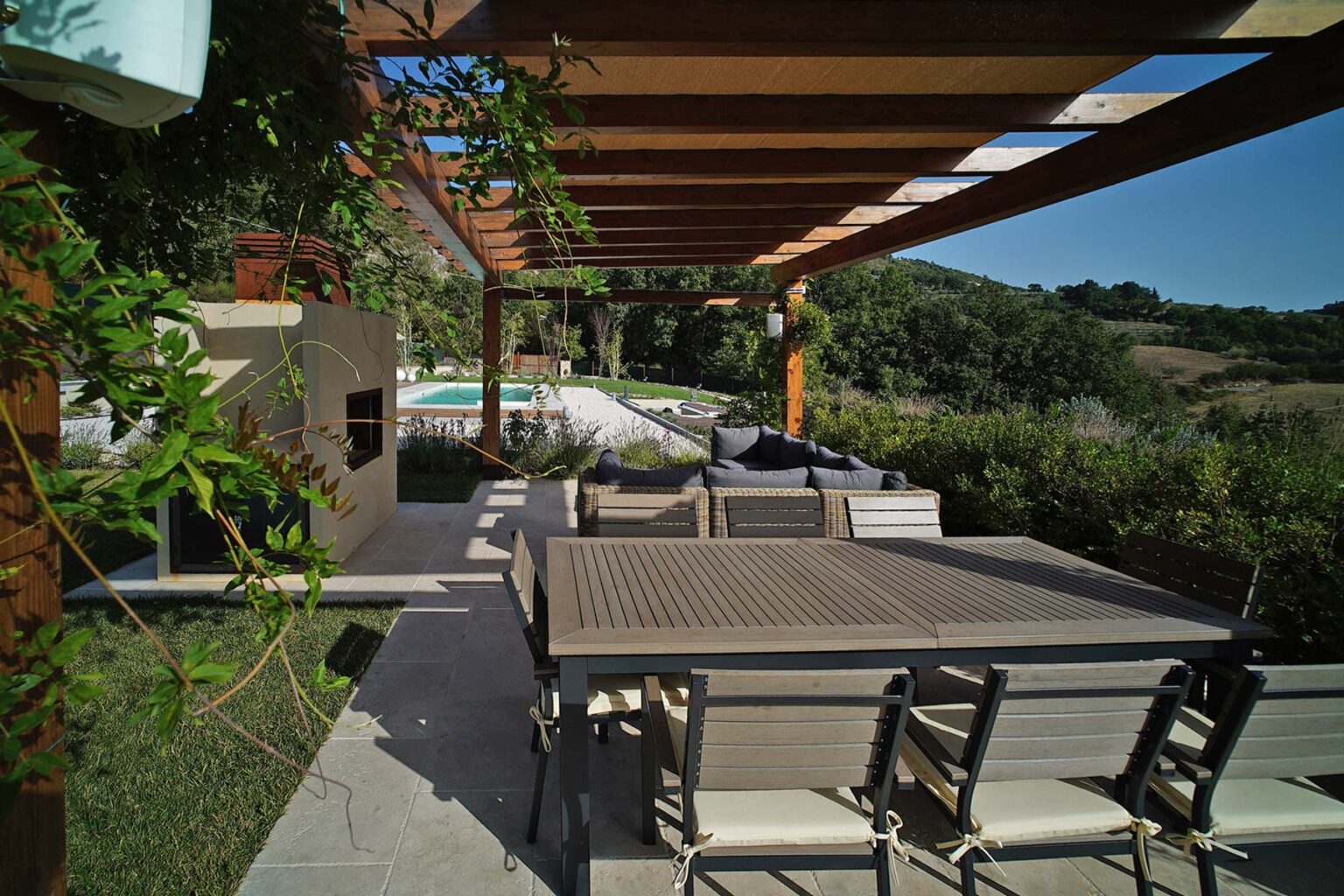 Modern outdoor patio with a wooden pergola, featuring a dining table and chairs, surrounded by greenery and a view of a pool in the background. Ideal for entertaining and relaxation. | Only on SopranoVillas