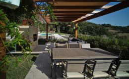 Modern outdoor patio with a wooden pergola, featuring a dining table and chairs, surrounded by greenery and a view of a pool in the background. Ideal for entertaining and relaxation.