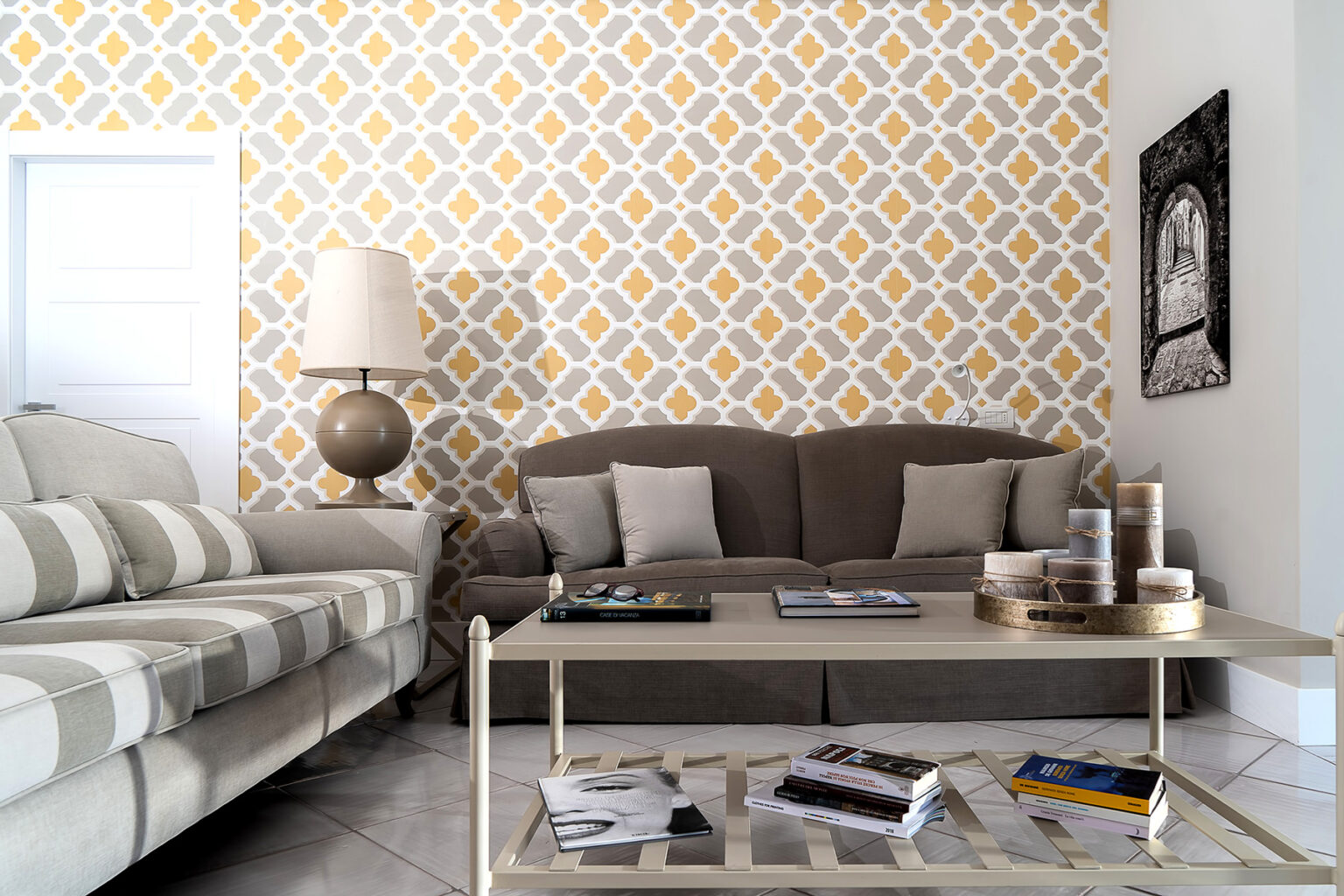 Modern living room with a patterned yellow and white accent wall, featuring a gray sofa, striped armchair, and a stylish coffee table with decorative items and magazines. | Only on SopranoVillas