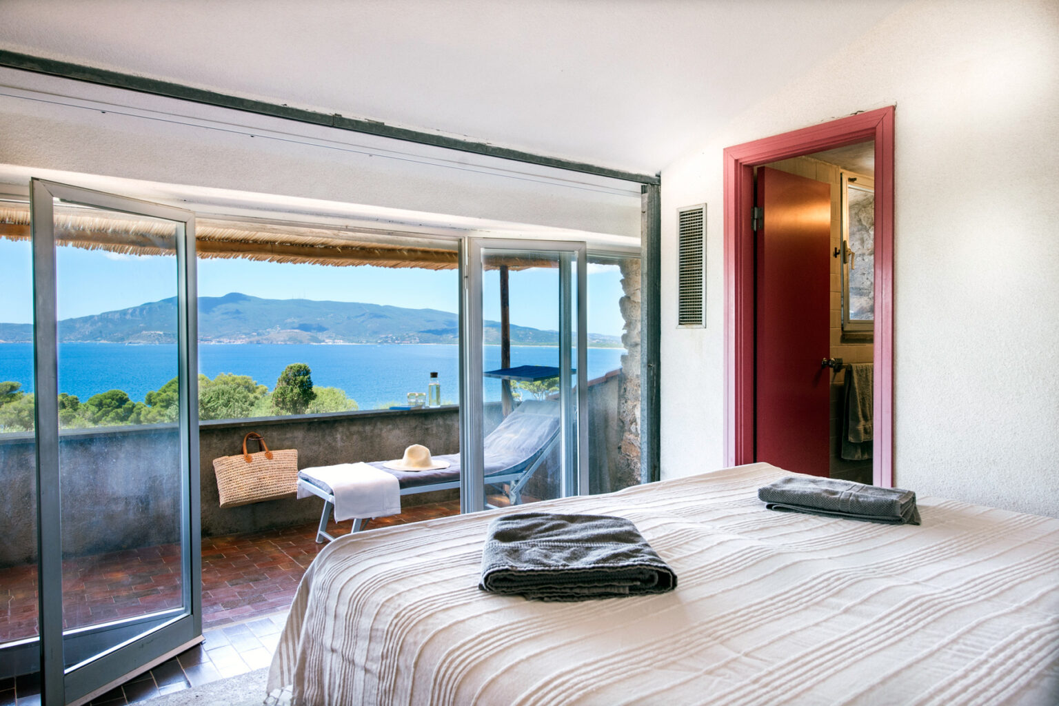Cozy bedroom with a stunning sea view, featuring large open windows, a comfortable bed with neatly arranged towels, and a relaxing outdoor seating area. Ideal for a tranquil getaway. | Only on SopranoVillas