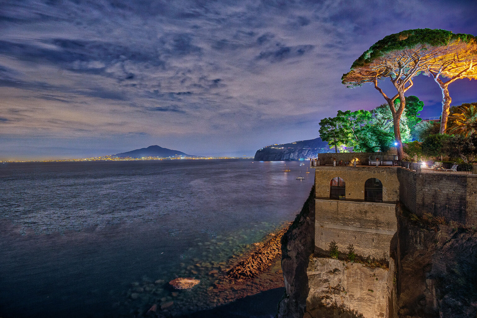 Scenic view of the coastline at night, featuring a cliff with lush trees, illuminated by soft lights, overlooking the calm sea and distant mountains. Ideal for showcasing the beauty of coastal landscapes and nighttime photography. | Only on SopranoVillas