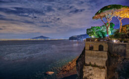 Scenic view of the coastline at night, featuring a cliff with lush trees, illuminated by soft lights, overlooking the calm sea and distant mountains. Ideal for showcasing the beauty of coastal landscapes and nighttime photography.